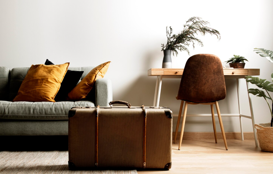Une valise vintage est posée au sol au centre d'un salon meublé avec soin, à côté d'un canapé gris aux coussins jaunes et d'un petit bureau en bois avec sa chaise marron